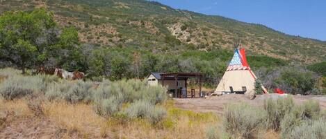 A traditional tipi with a red and blue design, a small wooden structure, and a hilly landscape in the background.