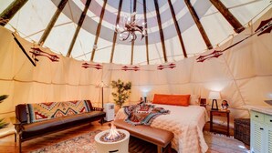 A cozy interior of a tent with a bed, a bench, a small table, and a fire pit.