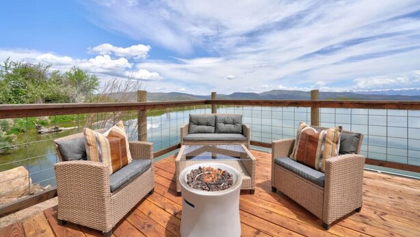 A deck with wicker furniture and a glass-top table, overlooking a body of water and distant mountains.