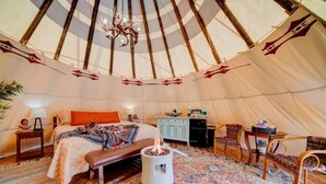 A cozy interior of a tent with a bed, a small table, a chair, and a fire pit.