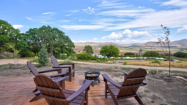 Una terraza de madera con cuatro sillas Adirondack y un brasero, con vistas a un paisaje pintoresco con árboles y colinas lejanas.