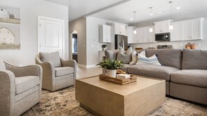 A modern living room with a beige sofa, two armchairs, and a wooden coffee table. There is a kitchen in the background with white cabinets and pendant lights. - 6 Bed 6 Bath Sleeps 22 w/4 Private Master Suites! (Hurricane)