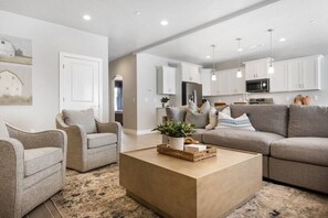 Een moderne woonkamer met een beige bank, twee fauteuils en een houten salontafel. Achtergrond is een keuken met witte kastjes en hanglampen.
