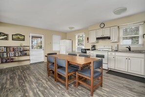 A compact kitchen with white cabinets, a refrigerator, a microwave, and a stove. There is a dining table with four chairs and a bookshelf filled with books. - Ryokan | Unobstructed Oyster Bay Waterfront Views w/ Screened Porch, Balconies & Boat Launch Access (Chincoteague Island)