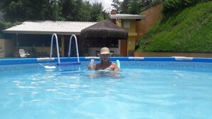 A swimming pool with a ladder, a person in the water, and a building with a thatched roof in the background. - Large house in a gated community, 3 bedrooms, 5 bathrooms and a large backyard. (Atibaia)