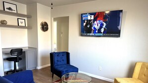 A living room with a blue sofa, a yellow armchair, a glass coffee table, and a TV displaying a sports game. - A Beautiful Storey - Quiet Condo minutes from Midland Memorial (MIDLAND)
