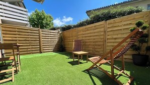 Un patio trasero con césped artificial verde, una cerca de madera, una silla de madera, una mesita y un parque infantil de madera.