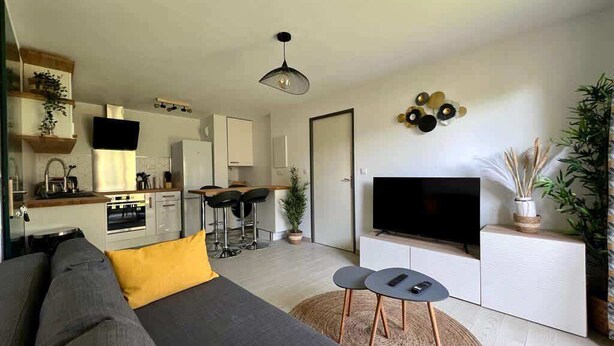 Una sala de estar moderna con sofá, televisión y comedor, con una cocina al fondo.