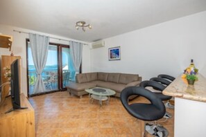 Una sala de estar con un sofá, una mesita redonda y un televisor sobre un mueble de madera. Hay un balcón con vistas al mar.