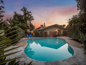 Une piscine avec un espace patio, entourée de verdure, et une maison avec un toit en pente.