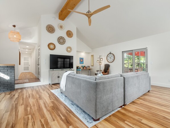 Un salon spacieux avec un canapé d’angle gris, un téléviseur à écran plat, un plancher en bois et un ventilateur de plafond.