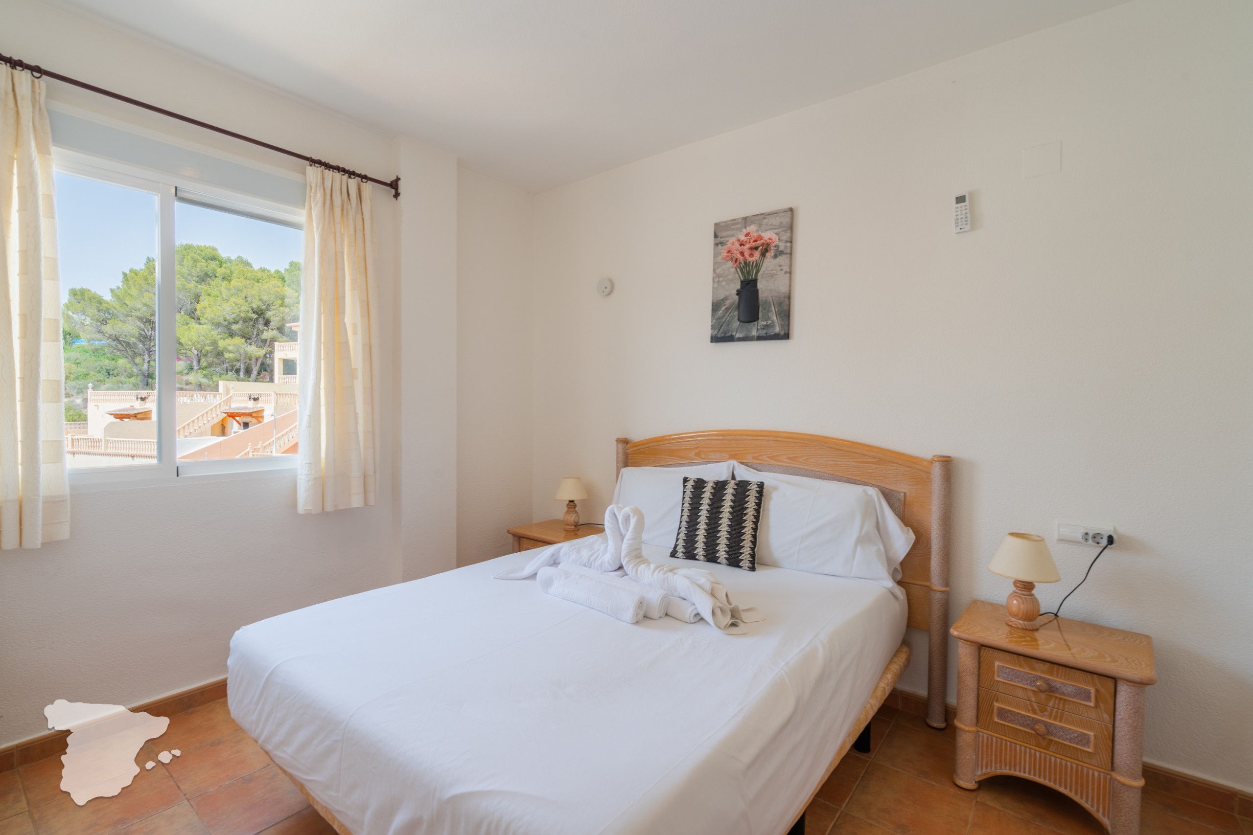 Quarto de hotel com cama, mesa de cabeceira, luminária, pintura e janela com cortinas.
