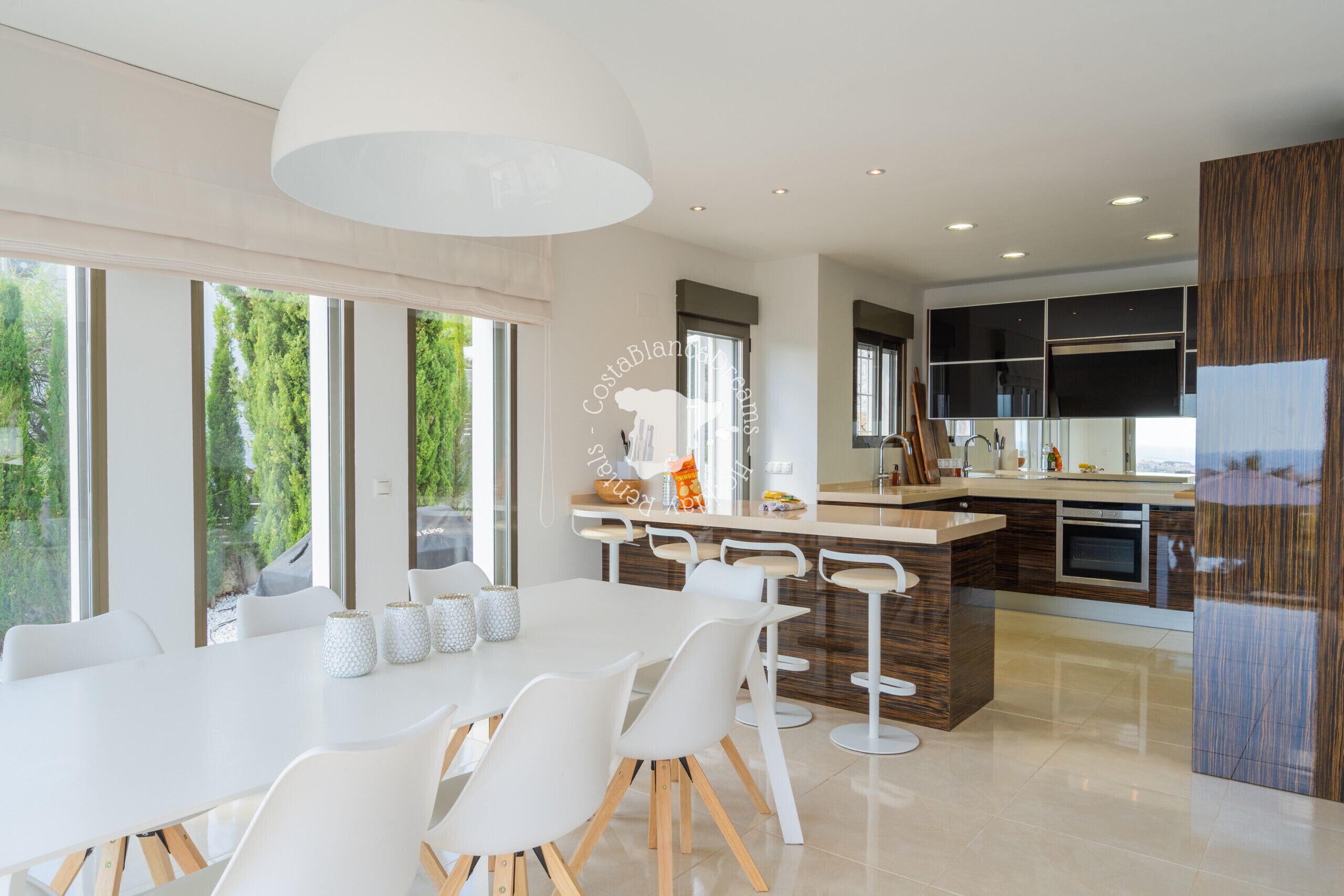 Una cocina moderna con una isla grande, taburetes blancos tipo bar y un área de comedor con mesa y sillas blancas.
