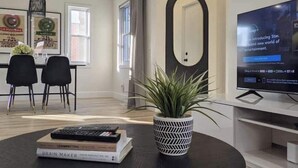 A modern living room with a dining area, a large mirror, a potted plant, and a television displaying a movie poster. - Perfect for large groups, lots of parking, 5BRs, Game Room, Fast Wifi (Kitchener)