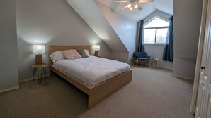 Ein Schlafzimmer mit schrägverdeckter Decke, einem Bett, zwei Nachttischlampen, einem Stuhl und einem fenster mit Vorhängen.