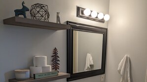 Ein Badezimmer mit Toilette, Waschbecken und Spiegel. Über dem Waschbecken befinden sich Regale mit Pflegeprodukten und Dekoration.