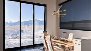 A dining area with a large table and chairs, a modern chandelier, and a view of a snowy landscape through large windows. - Modern 3 bedroom home on 20 private acres in Paradise Valley (Livingston)