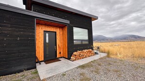 A modern log cabin with a black exterior, a wooden door, a window, and a stack of firewood outside. - Modern 3 bedroom home on 20 private acres in Paradise Valley (Livingston)