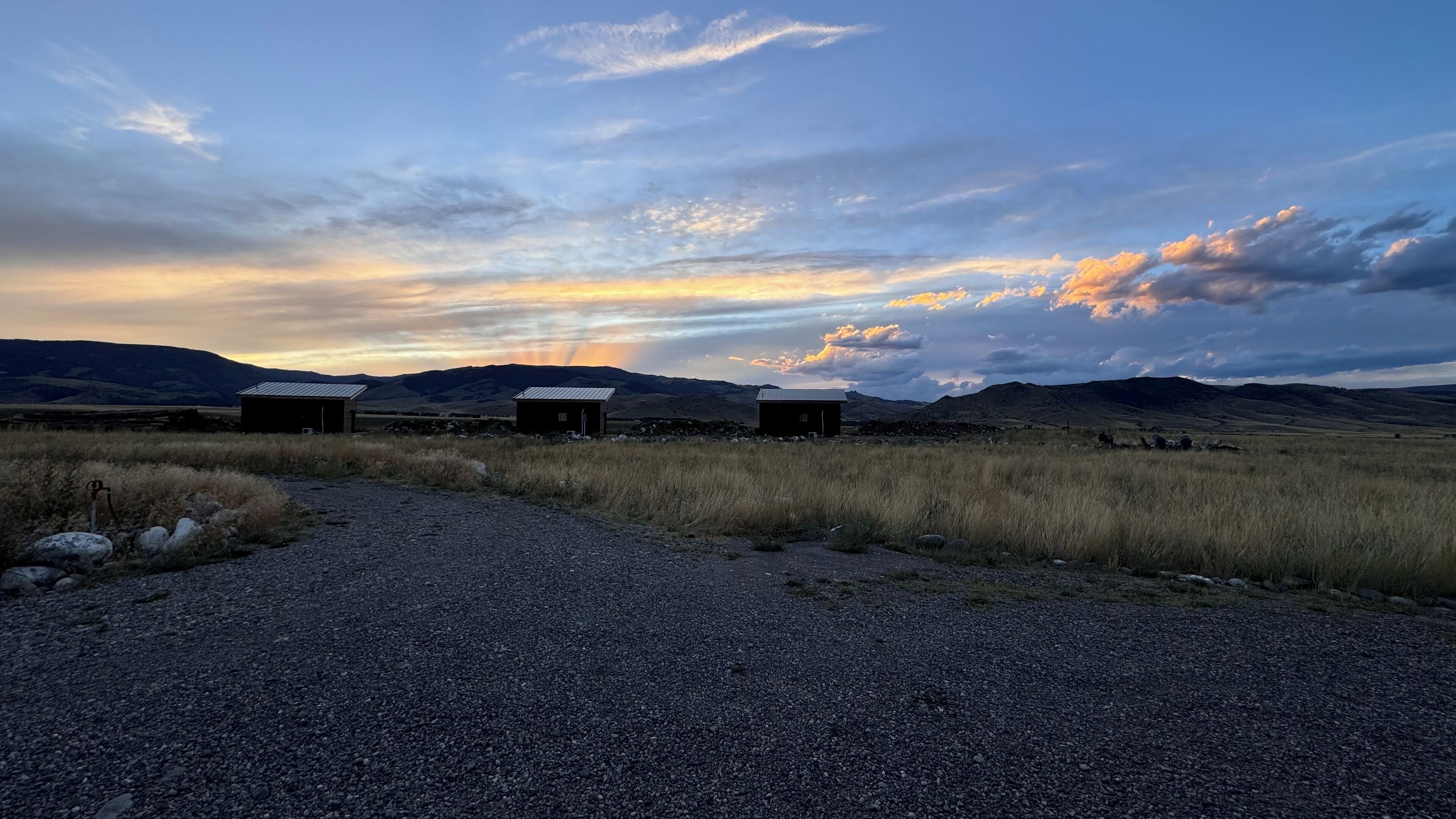Un chemin de gravier mène à trois petits bâtiments situés dans un cadre rural, avec une chaîne de montagnes en arrière-plan et un ciel au coucher du soleil.