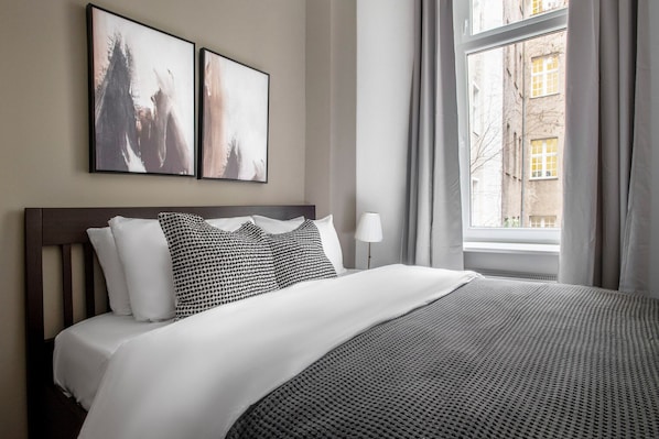 A neatly made bed with a white comforter and a patterned blanket, a wooden headboard, two wall-mounted abstract paintings, a bedside lamp, and a window with curtains.