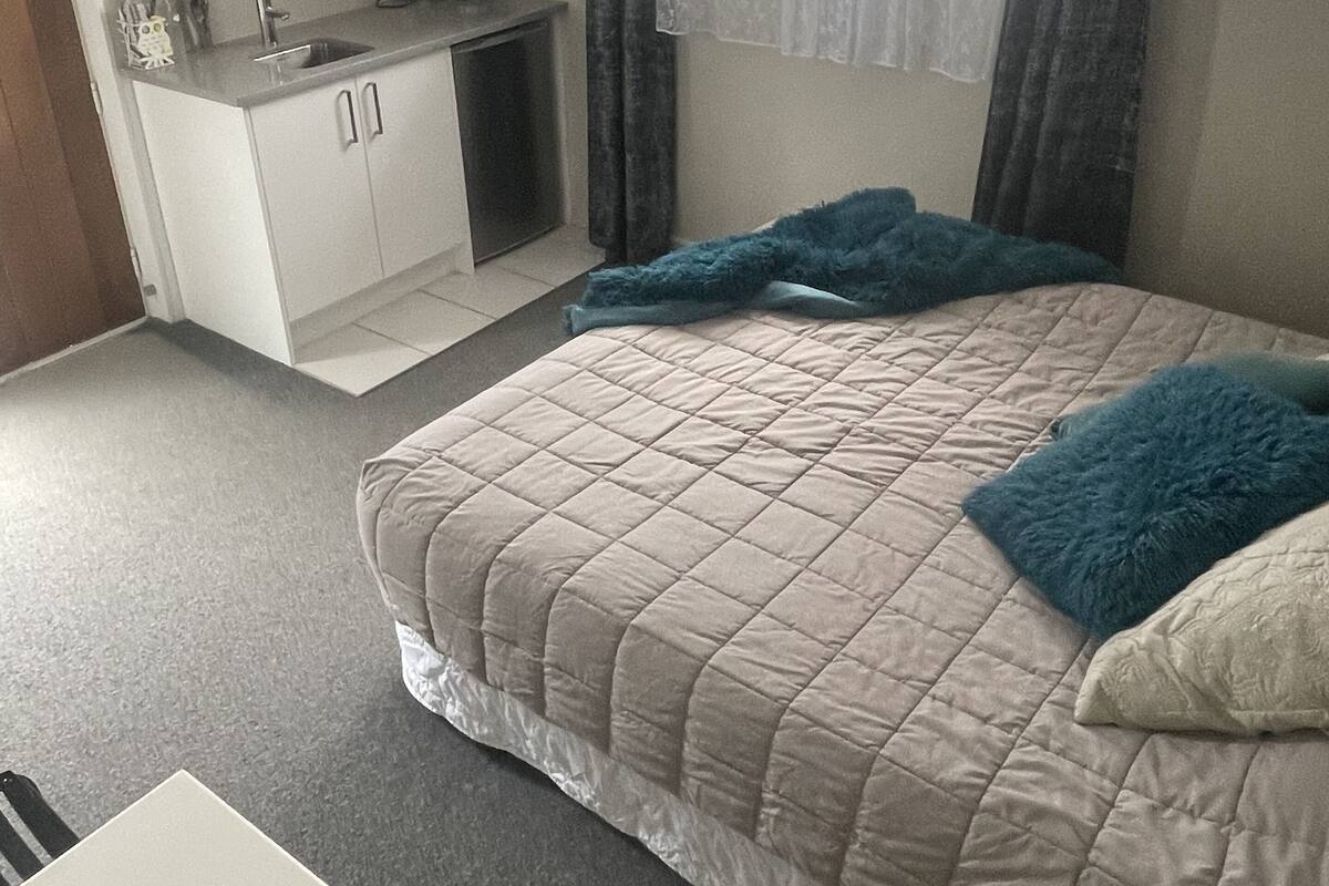 Quarto com cama, escrivaninha, pequeno espaço de cozinha e janela com cortinas.