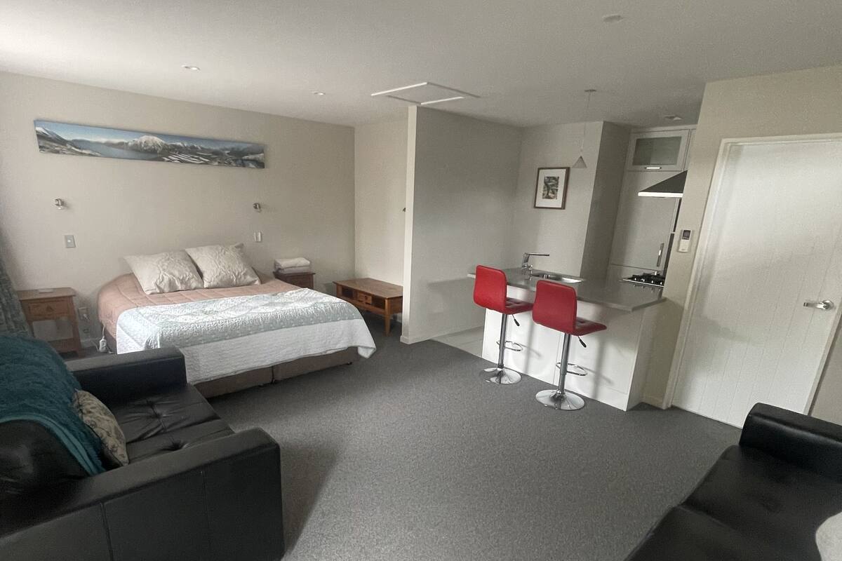 Quarto de hotel moderno com cama, sofá, pequena mesa e área de cozinha compacta.