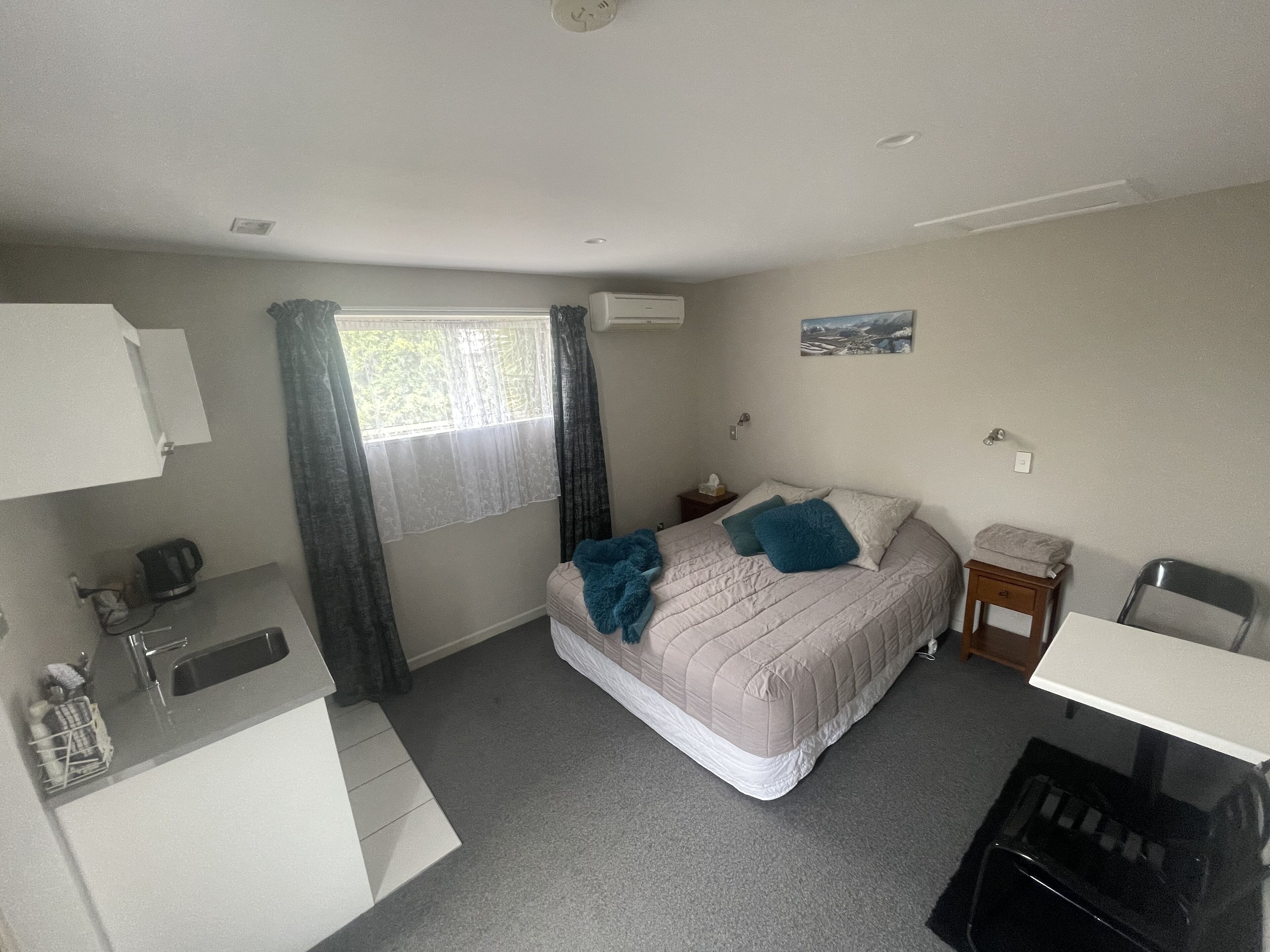 Ein kleines Hotelzimmer mit einem Bett, einem Schreibtisch, einem Stuhl, einem kleinen Tisch, einem Fenster mit Vorhängen, einer Wand-Klimaanlage und einem Wandbild.