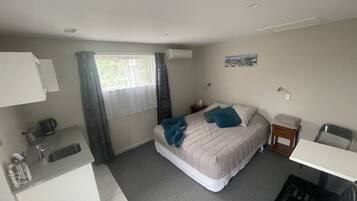 Ein kleines Hotelzimmer mit einem Bett, einem Schreibtisch, einem Stuhl, einem kleinen Tisch, einem Fenster mit Vorhängen, einer Wand-Klimaanlage und einem Wandbild.