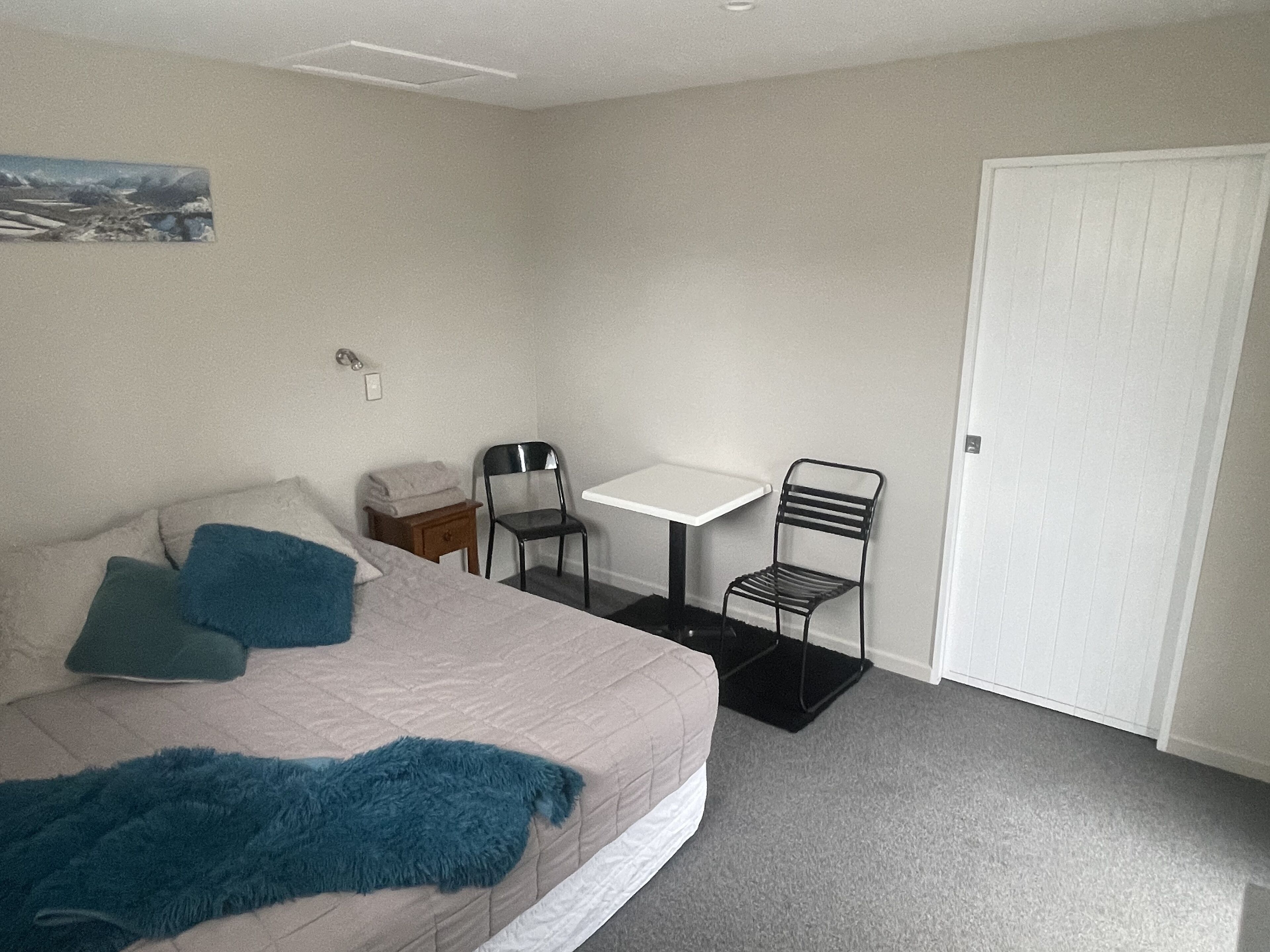 A bedroom with a bed, two chairs, a small table, a door, and a painting on the wall.