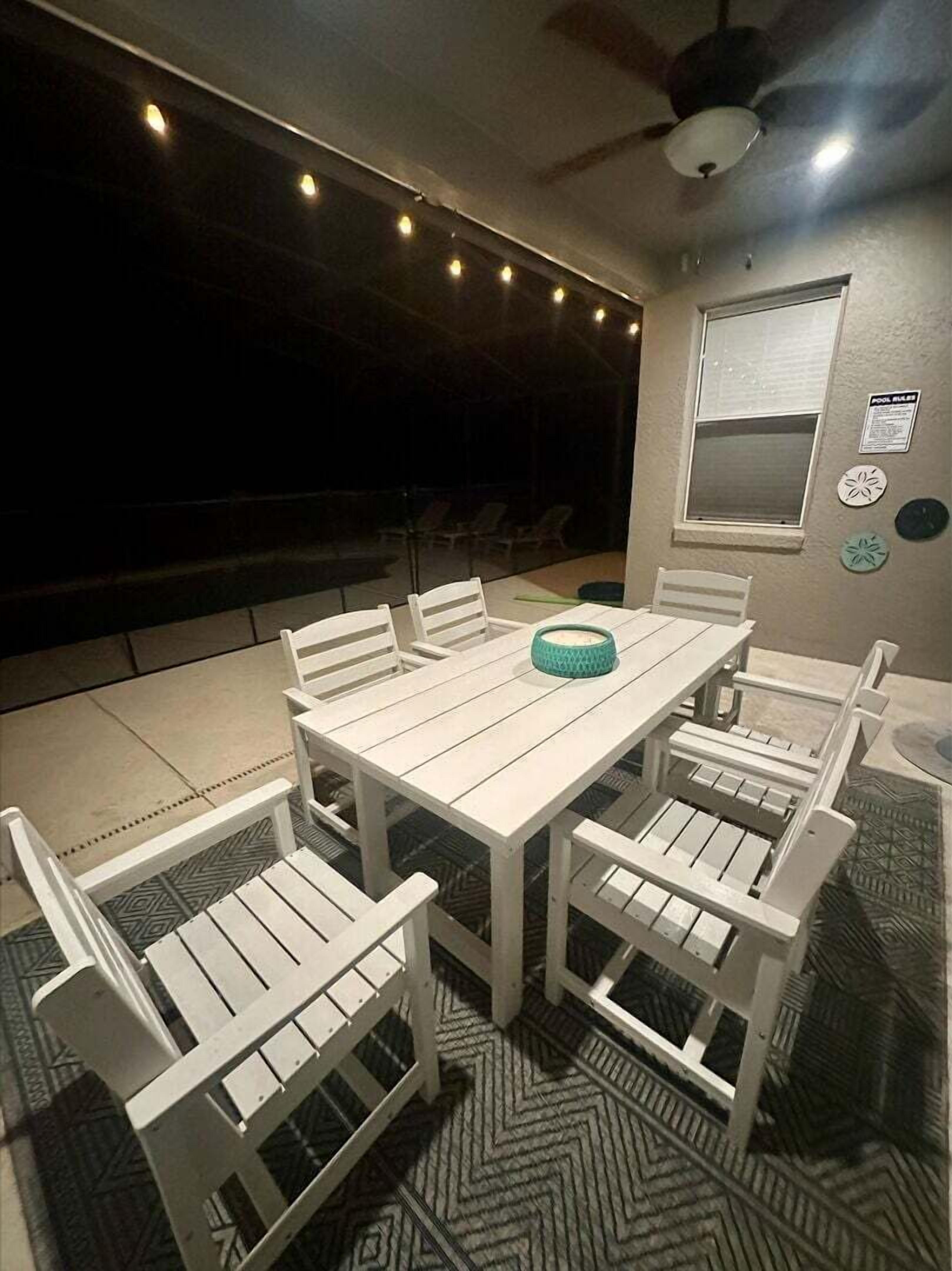 Un patio techado con una mesa y sillas blancas, un ventilador de techo y luces colgantes.