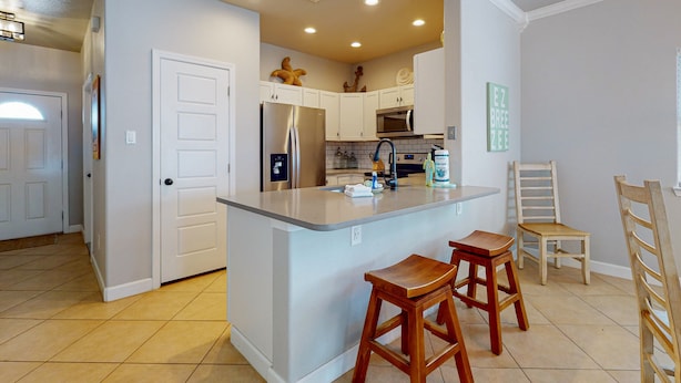 Una cocina con armarios blancos, una isla central con taburetes, un refrigerador y un microondas.