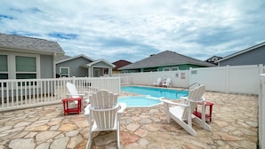 Un patio trasero con piscina, sillas Adirondack blancas y un patio de piedra.