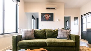 Een groene bank met geruite kussens in een woonkamer met een groot raam, een hangend fotolijstje en een zichtbare keuken door een open deur.
