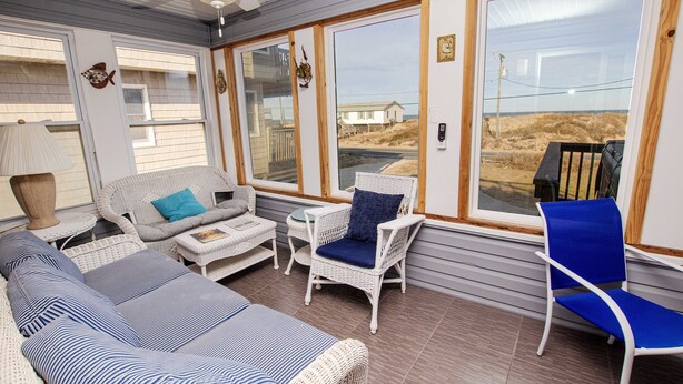 A covered outdoor seating area with wicker furniture, a ceiling fan, and large windows offering a view of the beach.