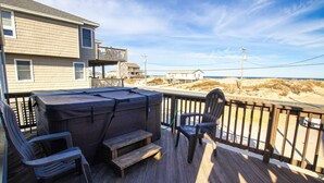 A wooden deck with a hot tub, chairs, and steps leading to a balcony overlooking a beach.