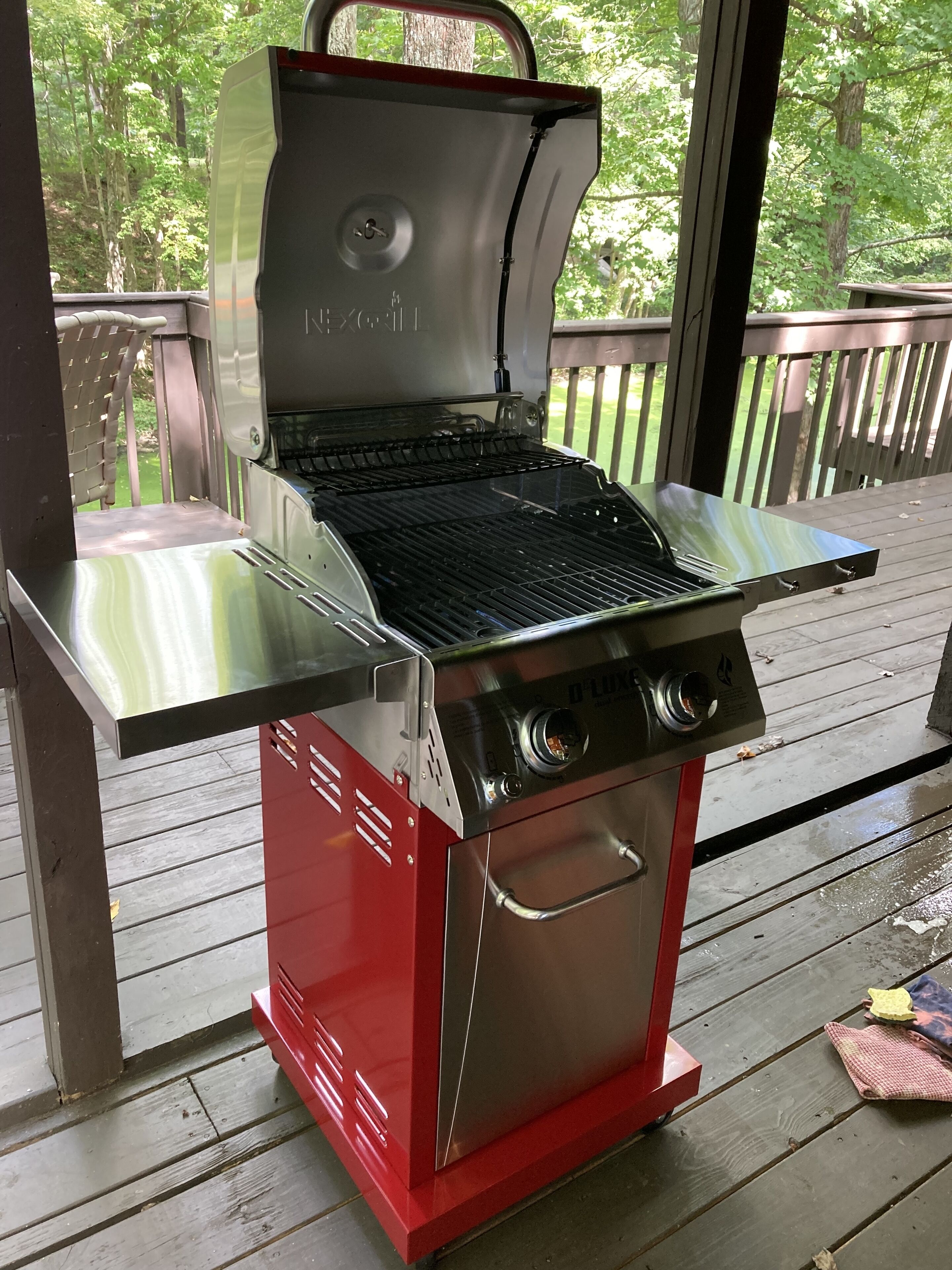 Una parrilla para barbacoa de acero inoxidable con base roja, ubicada en una terraza de madera con vista a un área boscosa.
