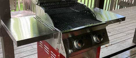 Una parrilla para barbacoa de acero inoxidable con base roja, ubicada en una terraza de madera con vista a un área boscosa.