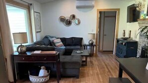 Un saló amb un sofà, una taula de centre i un moble per a la televisió. Hi ha una finestra amb cortines, una làmpada i aire condicionat fixat a la paret.