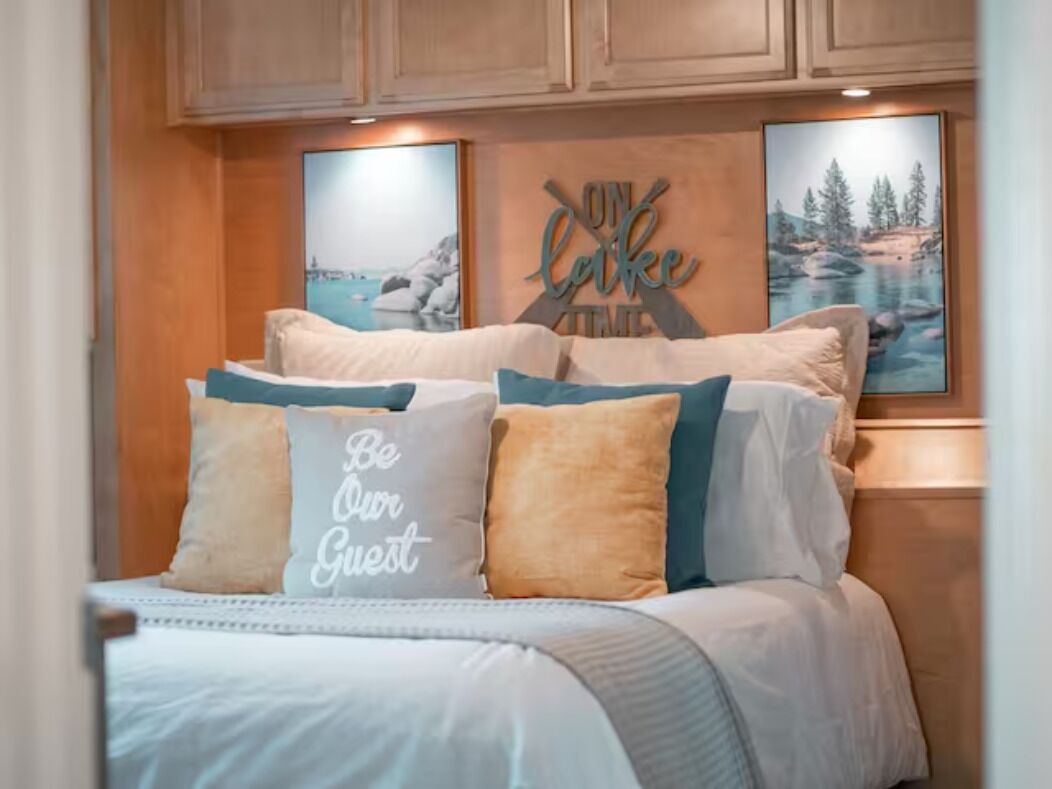 A bed with a 'Be Our Guest' pillow, wooden headboard, and two framed pictures on the wall.
