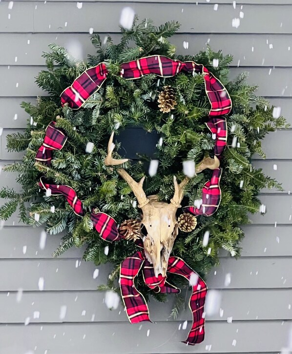 A Christmas wreath with a deer skull centerpiece, pine branches, and red plaid ribbons, hanging on a gray wooden wall.