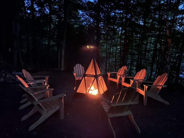 A campfire with a tent labeled "Firefly" surrounded by wooden chairs in a forest setting.