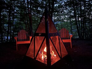 A campfire with a metal screen, surrounded by two wooden chairs, set in a forest.