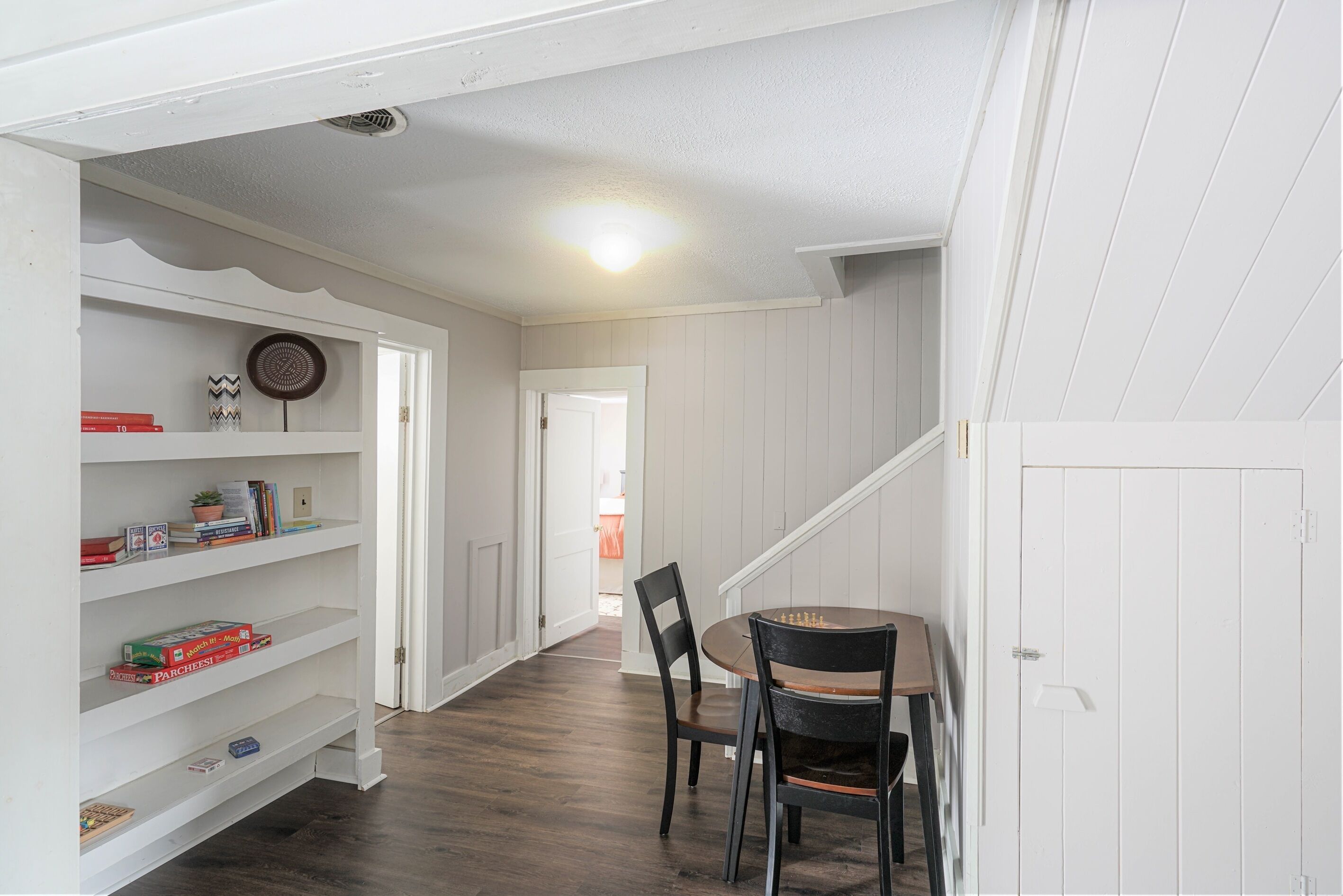 Un couloir comprenant un coin repas avec une table et des chaises, une bibliothèque remplie de livres et d’objets décoratifs, et une porte donnant sur une autre pièce.