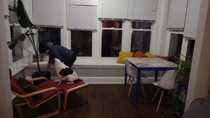 Une pièce chaleureuse avec un plancher en bois, un coin fenêtre agrémenté de fauteuils rouges, une table recouverte d’une nappe colorée et une plante en pot.