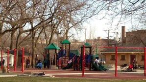 Ein Spielplatz mit verschiedenen Spielgeräten, darunter Rutschen und Kletterbereiche, umgeben von Bäumen und einem Gebäude im Hintergrund.