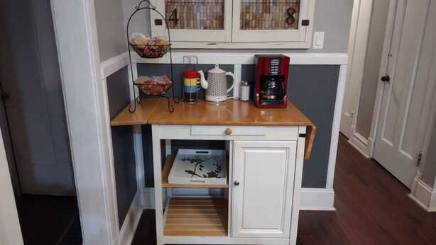 Un petit coin cuisine avec un plan de travail en bois, un meuble blanc doté d’un tiroir et une étagère contenant divers objets, notamment un panier, une tasse et une machine à café.
