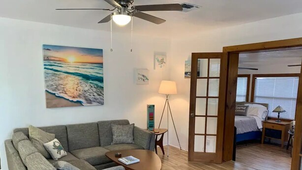 A modern living room with a grey sofa, two wooden side tables, a ceiling fan, and a painting of a sunset over the ocean.
