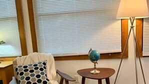 A room with a wooden floor, a beige armchair with a patterned pillow, a small round table with a globe, a floor lamp, and a window with blinds.