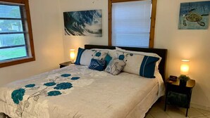 A bedroom with a bed, two nightstands, a painting of a wave, a painting of a turtle, and a window with blinds.