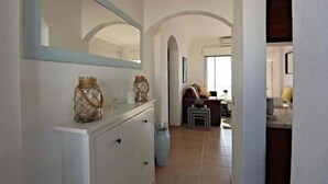 Un couloir avec une console blanche, un miroir et un vase. Une porte donne accès à une pièce où se trouvent un canapé et une petite table.